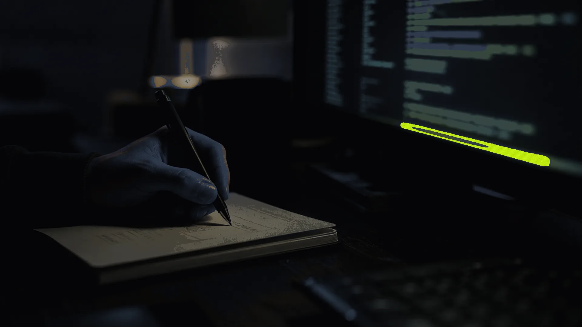 A hand mid-annotation in a notebook beside a monitor showing blurred code. Human and machine collaborating quietly.